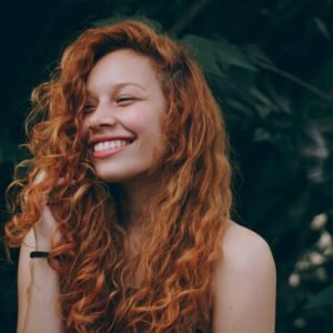 A happy woman with long, vibrant curly red hair smiling broadly while running her hand through her hair against a dark leafy background.